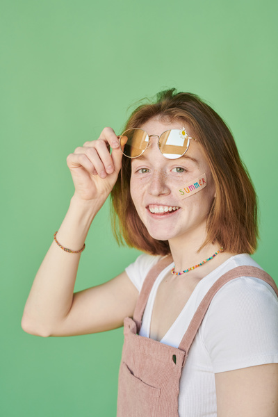 A smiling woman with red hair and a sticker with an inscription on her cheek lifting yellow sunglasses with a daisy