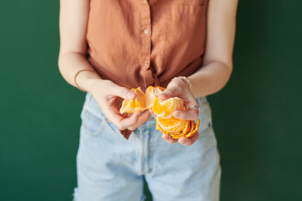 Close-up of a tangerine being cleaned by a woman in a sleeveless blouse and jeans standing on a green background