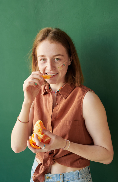 A woman with short red hair and a summer sticker on her face separating a segment from a peeled tangerine