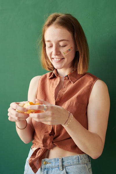 A woman with a red bob and a summer sticker on her face smiling holding a peeled tangerine in her hands