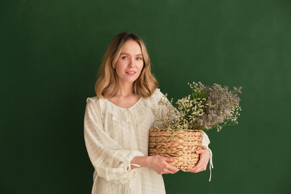 A woman with short blonde hair dressed in a white light dress holding a wicker basket with wildflowers in her hands