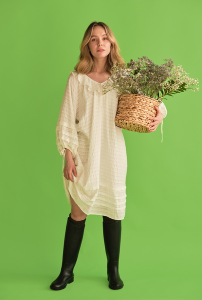 A woman with blonde hair dressed in a white summer dress and black boots holding the hem of her dress and a wicker basket with flowers and standing against a green background