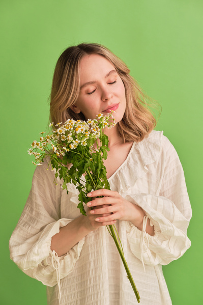 A woman with blonde hair dressed in white summer clothes with her eyes closed holding a bouquet of camomiles