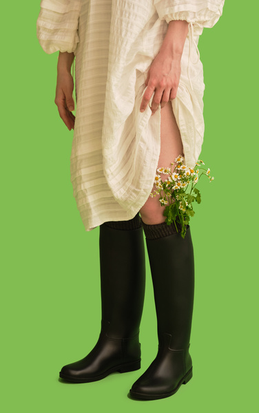 White daisies in a black boot of a woman lifting the hem of a white summer dress and standing against a green background