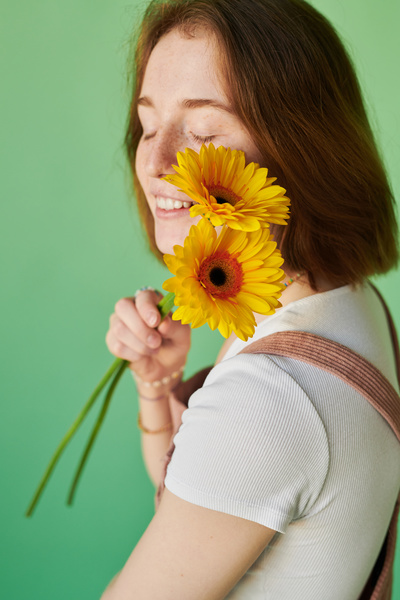A smiling red-haired woman with freckles holding yellow gerbera flowers near her face
