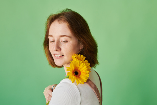 A smiling red-haired woman with freckles posing with yellow gerbera flowers on her shoulder