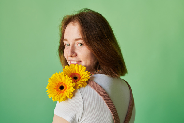 A redheaded woman with freckles smiles posing with yellow gerbera flowers on her shoulder