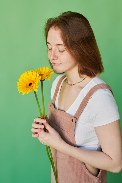 A woman with red hair and freckles dressed in a summer sundress smelling yellow gerbera flowers