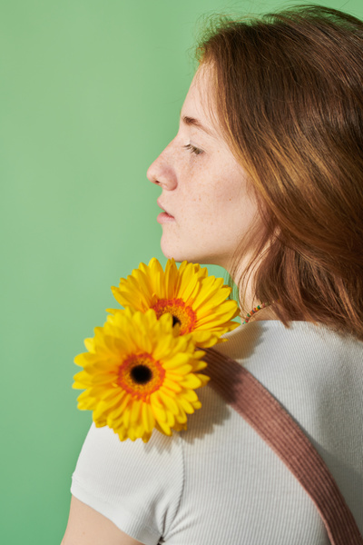A woman with red hair and freckles looking away holds yellow gerbera flowers on her shoulder