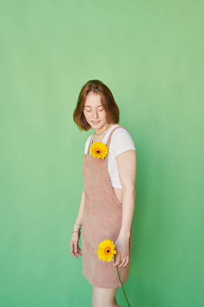A woman with red hair dressed in a summer pink corduroy sundress stands with a yellow gerbera in her hand with her head down