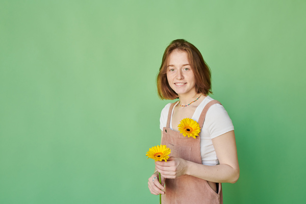 A woman with ginger hair dressed in a summer pink corduroy sundress standing with a yellow gerbera in her hand and smiling broadly