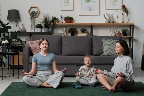 A dark haired mother and her children wearing light colored clothes meditating on the carpet in a livingroom