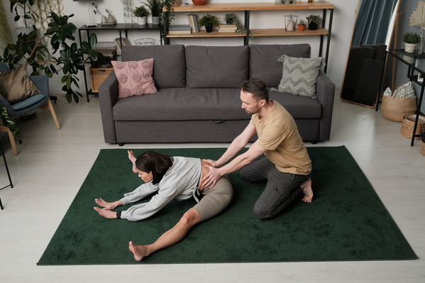 A man with a beard helping his wife with short hair to train stretching on a green carpet