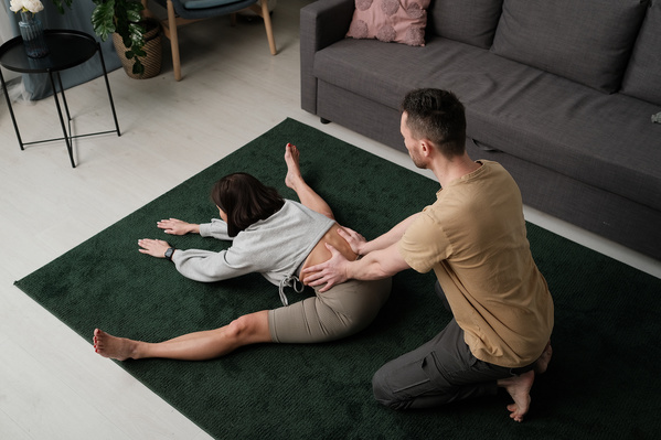 A man with a beard helping his wife with short hair to stretch to her feet on a green carpet
