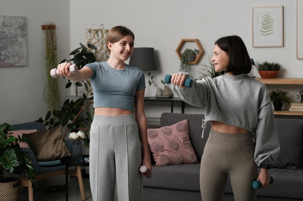 A teenage girl in light sports clothes and her mom with short dark hair doing an exercise with dumbbells