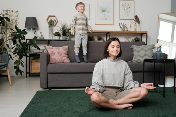 A woman with dark hair dressed in gray sportswear doing yoga while her son pampering on the couch