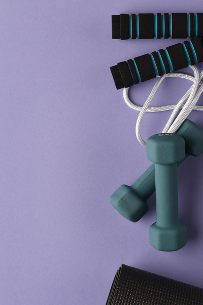 Top view of a skipping rope with black and blue handles laid on a purple surface with dumbbells and a rolled-up black yoga mat
