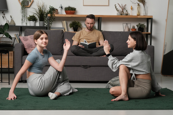 A teenage girl with tidied hair dressed in sports clothes and her dark-haired mother doing yoga while her father reads a book