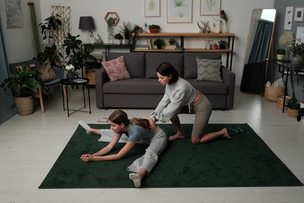 A woman with short dark hair dressed in a light sportswear helping her teenage daughter to do stretching in a living room