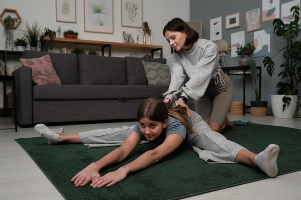 A beautiful woman with short dark hair dressed in a light sportswear helping her teenage daughter to perform stretching at home