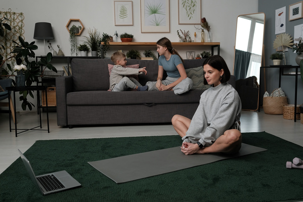 A woman with dark hair doing yoga online at home while her daughter communicates with her brother
