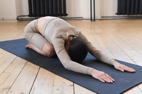 A woman with dark tidied up hair dressed in a beige sports outfit performing a balasana on a black sports mat