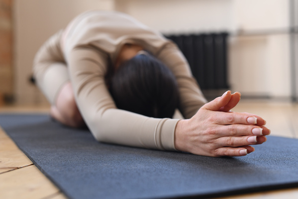 A woman with dark tidied up hair dressed in beige sportswear performing balasana on a black exercise mat