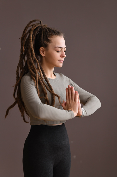 A woman with tidied up dreadlocks dressed in a dark-colored sports top closing her eyes performing anjali mudra