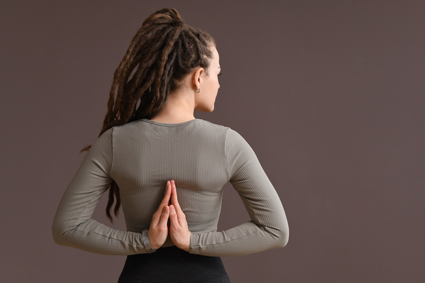 A woman with tidied up dreadlocks dressed in a dark-colored sports top folded her hands behind her back in a praying mudra