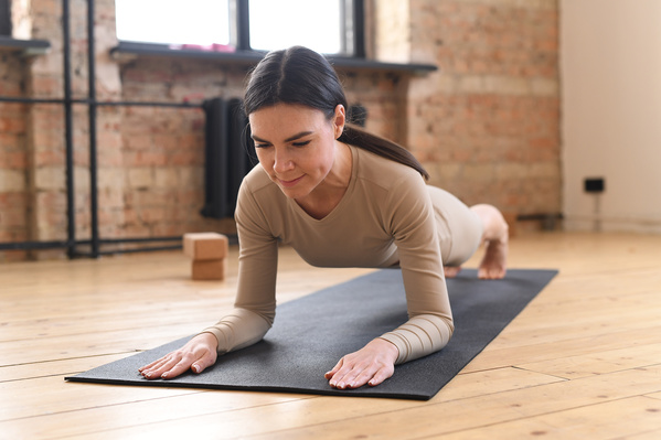 A woman with dark tidied up hair in beige sportswear performing a plank on a black sports mat
