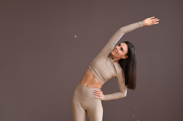 A woman with dark long hair dressed in a beige sports outfit performing a side bend against a beige background