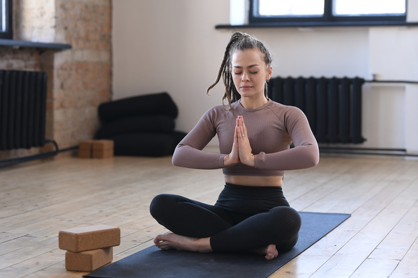 A young woman with tidied up dreadlocks dressed in gray sportswear meditating in the lotus position on a black sports mat in a loft room