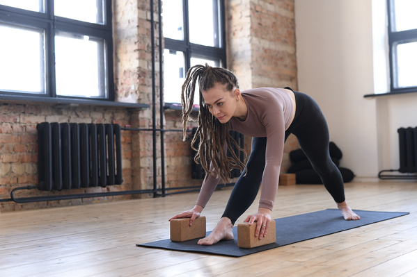 A beautiful woman with dreadlocks dressed in sportswear of dark shades doing leg stretching using yoga blocks on a black sports mat in a loft room