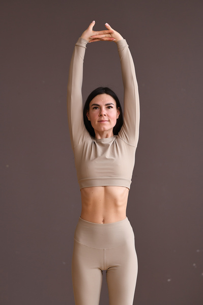 A woman with dark hair dressed in beige sports stretching her upper body against a beige background