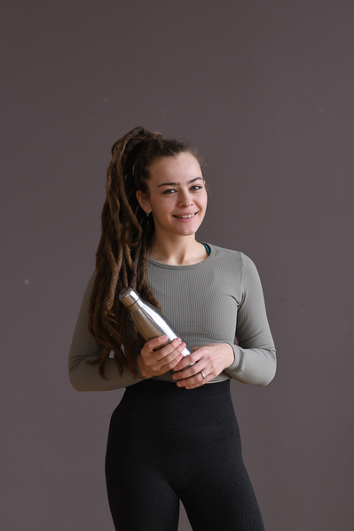 An athletic woman in a beige sports top and black leggings stands smiling with a metal water bottle