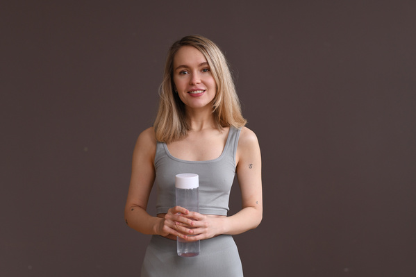 A sporty woman with blonde hair in a gray sports tight-fitting suit with a transparent water bottle on a beige background