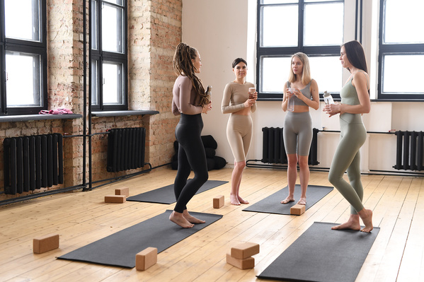Beautiful women dressed in sports clothes talking before a workout in the fitness hall with black yoga mats on the floor