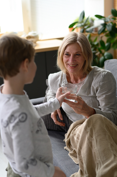 A blond boy in light clothes handing a glass of water to his grandmother lying on the sofa