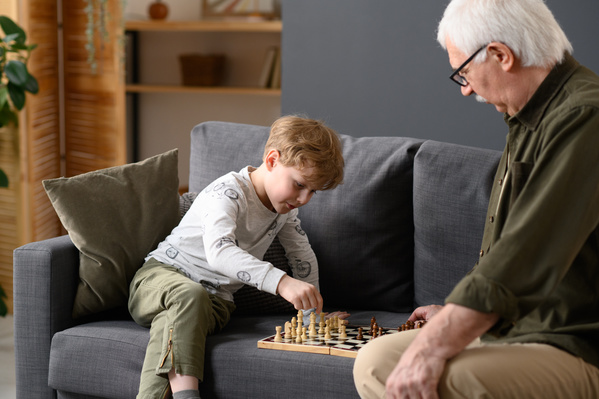 A little boy with blond hair playing chess with his grandfather on a gray sofa