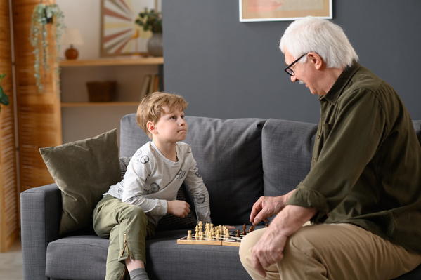 A little boy with blond hair and his grandfather playing chess on a gray sofa