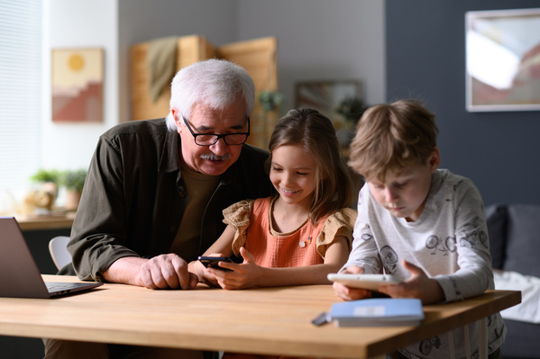 A gray-haired man in a dark shirt playing with his granddaughter a game on phone and a grandson using a tablet
