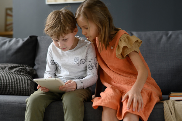 A girl with her hair gathered in a bright sundress watching her brother play a game on a tablet while sitting on the couch