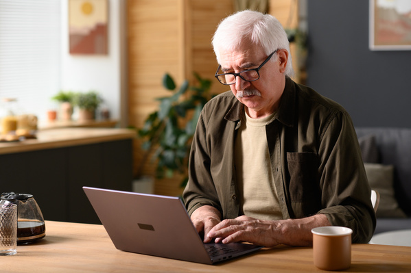 An elderly man in glasses working on a gray laptop at a wooden table at home