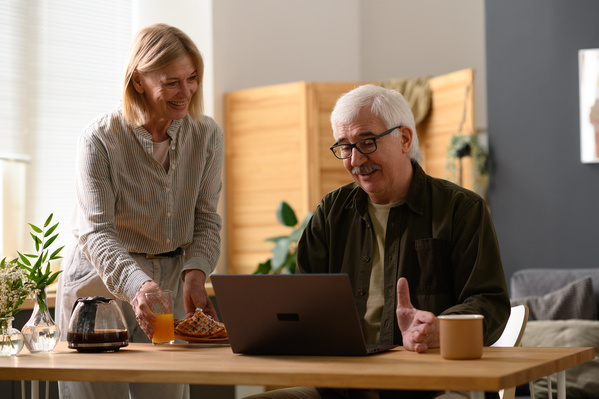An elderly man in glasses using a gray laptop at a wooden table at home and his wife giving him breakfast