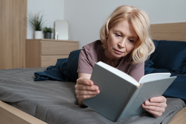 A senior woman with short blond hair in light house clothes reading in bed