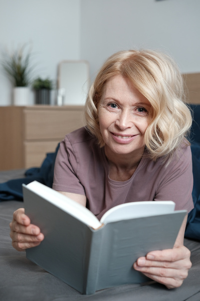 A smiling senior woman with short blond hair in light home clothes with a book in bed