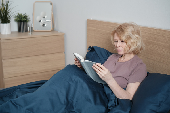 An aged woman with short blond hair in light house clothes reading a book in bed