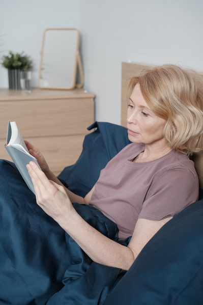 A senior woman with short blond hair in light house clothes reading a book in bed