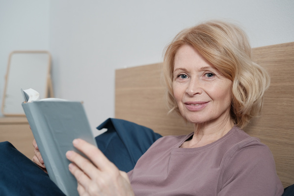 A smiling elderly woman with short blond hair in light home clothes with a book in bed
