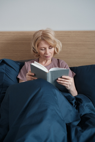 An elderly woman with short blond hair in light house clothes reading before going to sleep